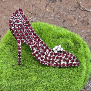 May include: A red rhinestone high heel shoe brooch. The shoe is made of silver metal and is covered in red rhinestones. The shoe is on a green mossy surface.