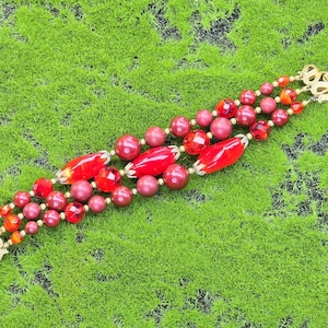 Op de afbeelding: Een vintage armband met drie strengen rode kralen in verschillende maten en vormen. De armband heeft ronde, ovale en facetgeslepen kralen in rode en bordeaux tinten. De sluiting en decoratieve elementen zijn goudkleurig.