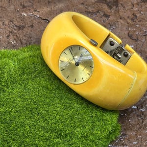 May include: A yellow plastic bangle watch with a round face and black hands. The watch has a metal clasp and is on a green mossy surface.