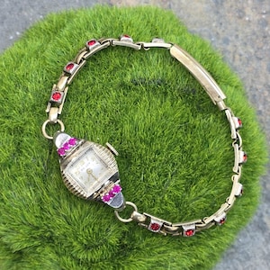 Puede incluir: Un reloj de pulsera de oro con una esfera cuadrada y una banda con joyas rojas.