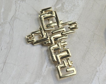 May include: Gold-toned cross pendant with a geometric design. The cross features a series of interconnected rectangular shapes, creating a modern and abstract aesthetic. The pendant is displayed on a light-colored, textured surface.