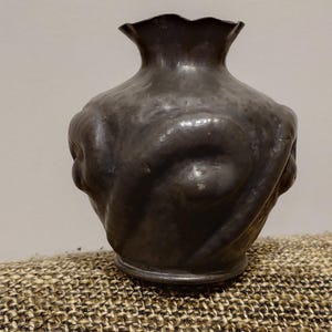 Può includere: Un vaso grigio scuro con un bordo a balze e un corpo bulboso. Il vaso ha una superficie strutturata con motivi a vortice e una piccola base circolare. Il vaso è in metallo e poggia su una superficie tessuta strutturata.