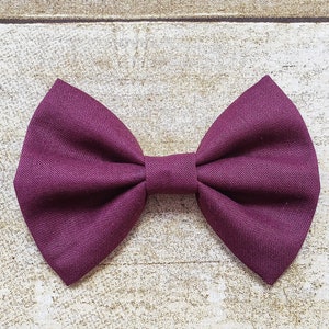 May include: A burgundy bow tie made of fabric. The bow tie is on a light brown wooden surface.