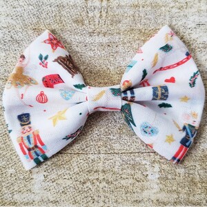 May include: A white bow with a Nutcracker-themed print. The bow features a variety of festive images, including nutcrackers, ballerinas, and Christmas ornaments. The bow is perfect for adding a touch of holiday cheer to any outfit.