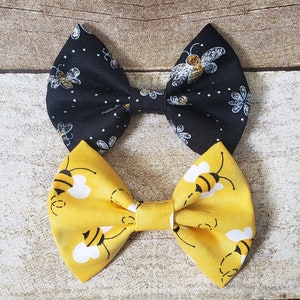 May include: Two fabric hair bows with a bee theme. One bow is black with white bees and white dots. The other bow is yellow with black bees and white clouds.