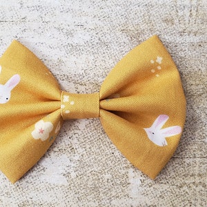 May include: A yellow bow with a white bunny rabbit print and small white flowers. The bow is made of fabric and has a simple design.