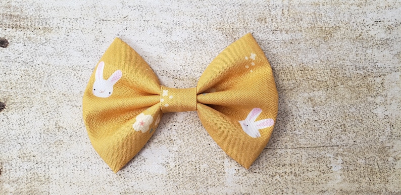 May include: A yellow bow with a white bunny rabbit print and small white flowers. The bow is made of fabric and has a simple design.