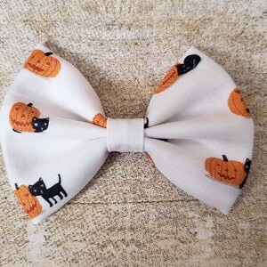 May include: A white fabric bow with a pumpkin and black cat pattern. The bow is tied in a classic bow shape.
