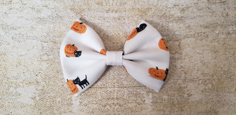 May include: A white fabric bow with a pumpkin and black cat pattern. The bow is tied in a classic bow shape.