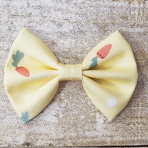 May include: A yellow fabric bow with a carrot print. The bow is tied in a classic bow shape and has a white center.