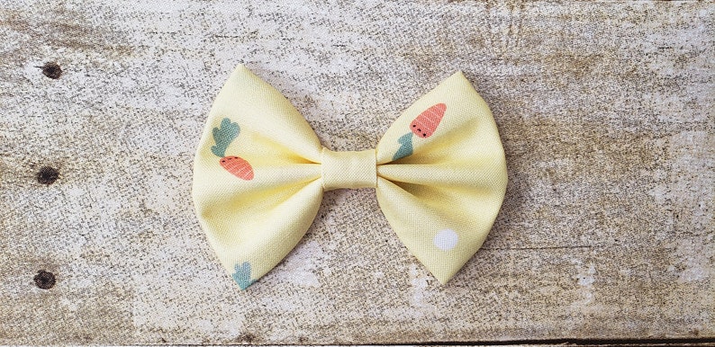 May include: A yellow fabric bow with a carrot print. The bow is tied in a classic bow shape and has a white center.