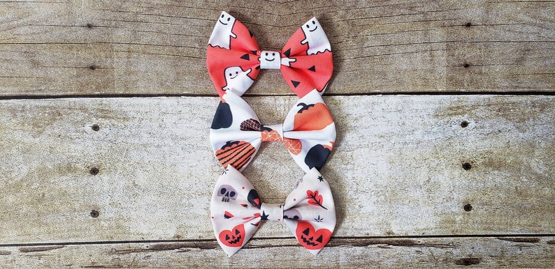 Puede incluir: Tres pajaritas con tem&aacute;tica de Halloween con diferentes dise&ntilde;os. La pajarita superior es naranja con fantasmas blancos. La del medio tiene calabazas y murci&eacute;lagos. La inferior tiene calaveras y calabazas. Todas sobre fondo de madera.