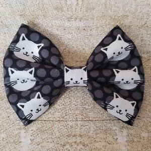 May include: A black and white bow with a polka dot pattern and white cat faces. The bow is made of fabric and has a loop on the back for attaching to hair.
