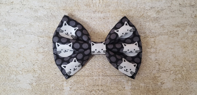May include: A black and white bow with a polka dot pattern and white cat faces. The bow is made of fabric and has a loop on the back for attaching to hair.