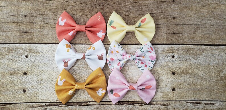 May include: Six fabric hair bows with different patterns. The bows feature bunnies, carrots, and floral designs in shades of pink, yellow, orange, and white.