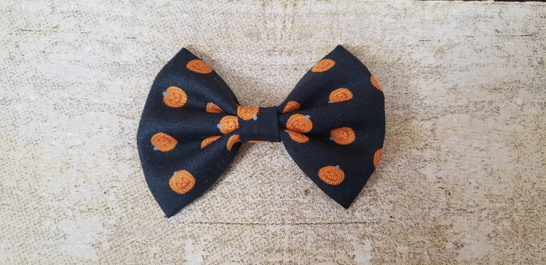 May include: A black bow tie with a pumpkin pattern. The bow tie is made of fabric and has a small loop on the back for attaching it to clothing.