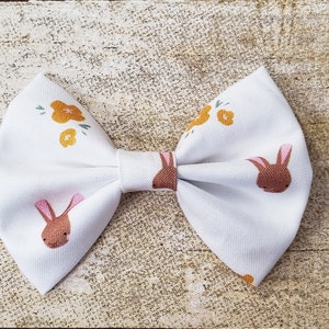 May include: A white bow with a brown bunny and yellow flower pattern. The bow is tied and ready to be worn.