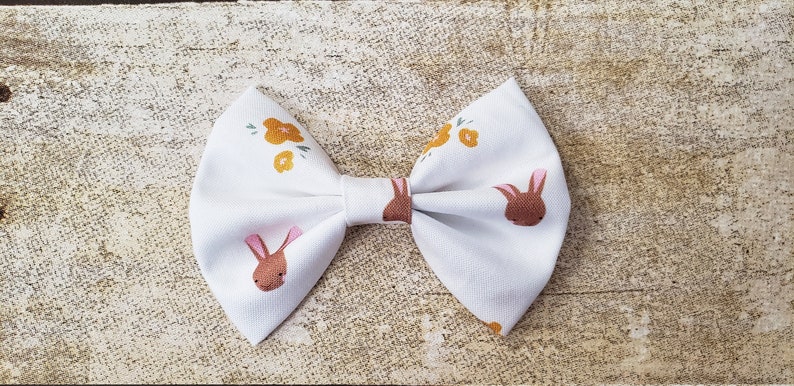 May include: A white bow with a brown bunny and yellow flower pattern. The bow is tied and ready to be worn.