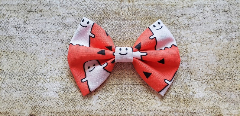Puede incluir: Una pajarita de Halloween hecha a mano. La pajarita presenta un patr&oacute;n repetido de fantasmas blancos con caras sonrientes sobre un fondo naranja brillante. Tambi&eacute;n hay tri&aacute;ngulos negros dispersos por el dise&ntilde;o.
