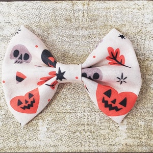 Puede incluir: Una pajarita de tem&aacute;tica Halloween de color crema con un estampado repetido de calaveras, corazones de calabaza y hojas de oto&ntilde;o. La pajarita est&aacute; atada en el centro con un nudo negro y detalles en forma de estrella.