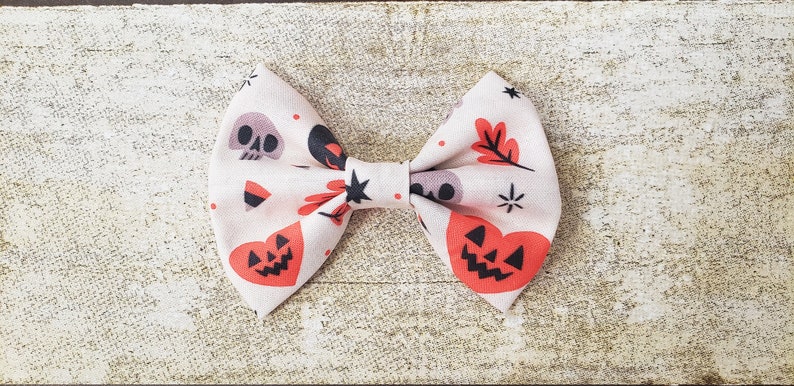 Puede incluir: Una pajarita de tem&aacute;tica Halloween de color crema con un estampado repetido de calaveras, corazones de calabaza y hojas de oto&ntilde;o. La pajarita est&aacute; atada en el centro con un nudo negro y detalles en forma de estrella.