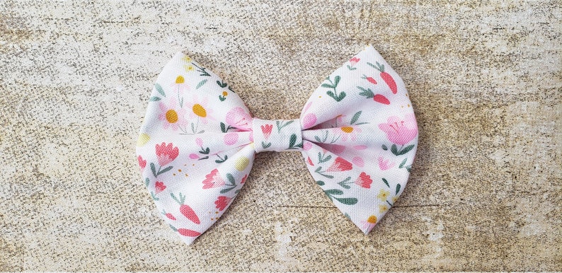 May include: A white fabric bow with a pink and green floral pattern. The bow is tied in a classic shape with a center loop.