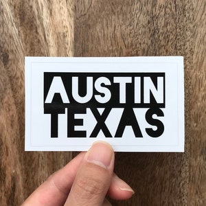 May include: A white rectangular sticker with the words "AUSTIN TEXAS" in bold, black capital letters. The sticker is held by a hand against a wooden background.