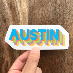 May include: A rectangular sticker with the word "AUSTIN" in layered, colourful text. The text is in shades of blue, orange, green, purple, and yellow. The sticker has a white border and is set against a wooden background.