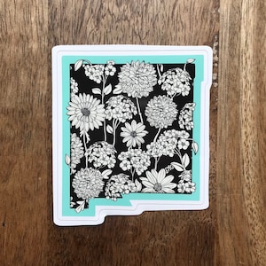 May include: A sticker featuring a floral design in black and white, set against a black background. The sticker has a turquoise border and is shaped like a state. The sticker is on a wooden surface.