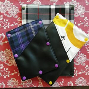 May include: Three fabric pouches with different patterns. The pouches are black, grey and white plaid, and white with a yellow abstract design. The pouches have purple and yellow snap closures.