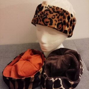 May include: A mannequin head is wearing a leopard print headband. Two additional animal print hats are displayed below. One hat has a zebra print exterior with an orange interior. The other hat has a giraffe print.