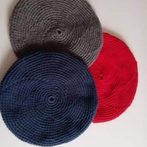 May include: Three crocheted berets in different colors: dark gray, navy blue, and red. Each beret has a circular design with a small hole at the center. The berets are displayed on a white background.