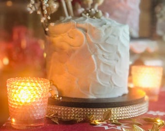 Puede incluir: Un pastel blanco glaseado con una decoración floral en la parte superior, sentado sobre un soporte para pastel de brillo dorado. El pastel está sobre un mantel rojo con una guirnalda de hojas dorées.