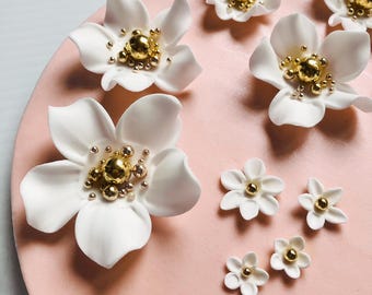 Op de afbeelding: Witte bloemvormige versieringen met gouden kralen in het midden zijn gerangschikt op een roze oppervlak. De bloemen variëren in grootte, met grotere op de voorgrond en kleinere op de achtergrond. De bloemblaadjes zijn glad en de gouden kralen voegen een vleugje elegantie toe.
