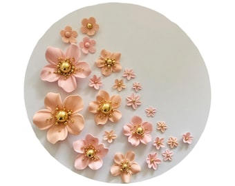 Può includere: Una collezione di decorazioni floreali rosa e pesca con centri dorati disposte su una superficie circolare bianca. I fiori variano di dimensioni, creando una composizione floreale decorativa. I fiori sono probabilmente fatti di un materiale morbido.