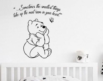 May include: Black and white Winnie the Pooh wall decal with a quote that reads "Sometimes the smallest things take up the most room in your heart."  The decal is on a white wall above a white cot with a white and grey patterned pillow.
