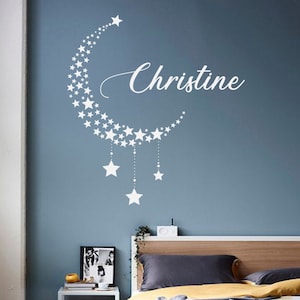 May include: White vinyl wall decal of a crescent moon with stars and the name Christine. The decal is on a blue wall above a bed with white bedding.