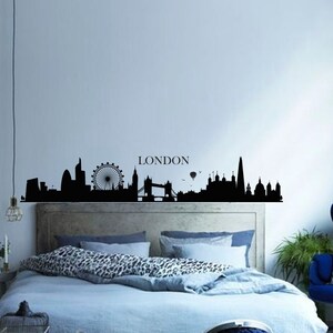 May include: Black and white silhouette of the London skyline with the London Eye, Big Ben, and Tower Bridge. The skyline is on a wall above a bed with blue bedding and a leopard print throw pillow.