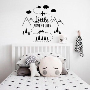 May include: Black and white wall decal featuring the text "Little Adventurer" and a design of mountains, trees, a car, and a plane. The decal is on a white wall above a white cot with white and black star patterned bedding.