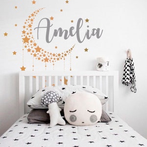 May include: A white cot with white and black star patterned bedding. A gold crescent moon with gold stars is on the wall above the cot. The name Amelia is written in grey cursive letters above the moon.