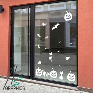 Puede incluir: Un escaparate de tienda decorado con calcomanías de temática de Halloween. Las calcomanías presentan calabazas, murciélagos, gatos y arañas. El escaparate está hecho de vidrio y tiene un marco negro.