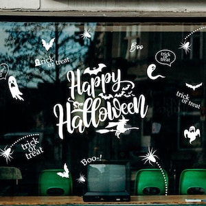 Puede incluir: Un escaparate de tienda decorado con pegatinas de temática de Halloween. Las pegatinas representan fantasmas, murciélagos, arañas y las palabras "Happy Halloween", "Boo!" y "Trick or Treat".