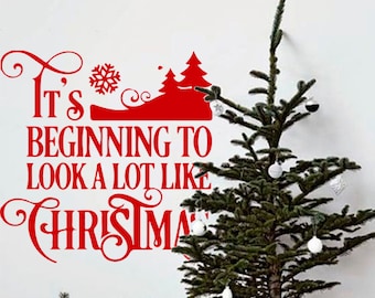 以下が含まれることがあります： 雪の結晶とスタイリッシュなクリスマスツリー2本が描かれた「It's beginning to look a lot like Christmas」というテキストの赤いビニール製のウォールステッカー。ステッカーは、白い壁に、小さな鉢植えのクリスマスツリーと、バスケットに入ったラッピングされたギフトと一緒に飾られています。
