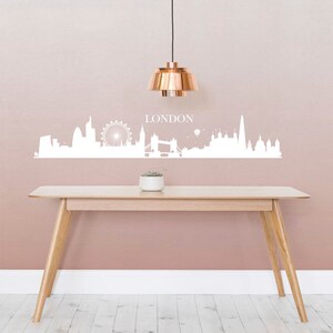 May include: White London skyline wall decal with the word "LONDON" above the skyline. The decal is on a light pink wall with a copper pendant light hanging above a wooden table.