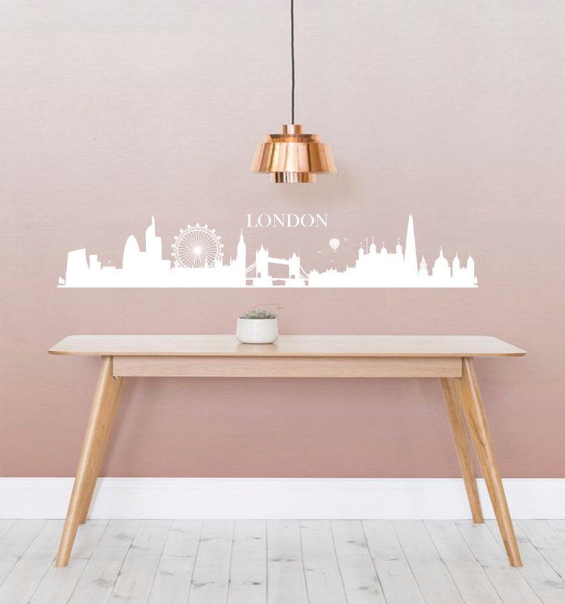 May include: White London skyline wall decal with the word "LONDON" above the skyline. The decal is on a light pink wall with a copper pendant light hanging above a wooden table.