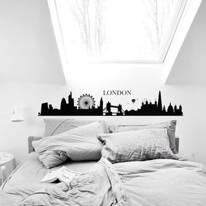May include: Black and white silhouette of the London skyline with the London Eye, Big Ben, and Tower Bridge. The skyline is on a white wall above a gray bed with pillows.