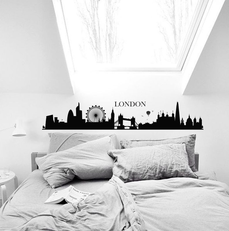May include: Black and white silhouette of the London skyline with the London Eye, Big Ben, and Tower Bridge. The skyline is on a white wall above a gray bed with pillows.