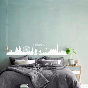 May include: A white silhouette of the London skyline with the word "LONDON" above it, on a light blue wall above a gray bed with white pillows.