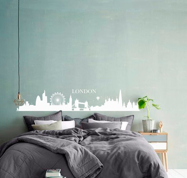 May include: A white silhouette of the London skyline with the word "LONDON" above it, on a light blue wall above a gray bed with white pillows.