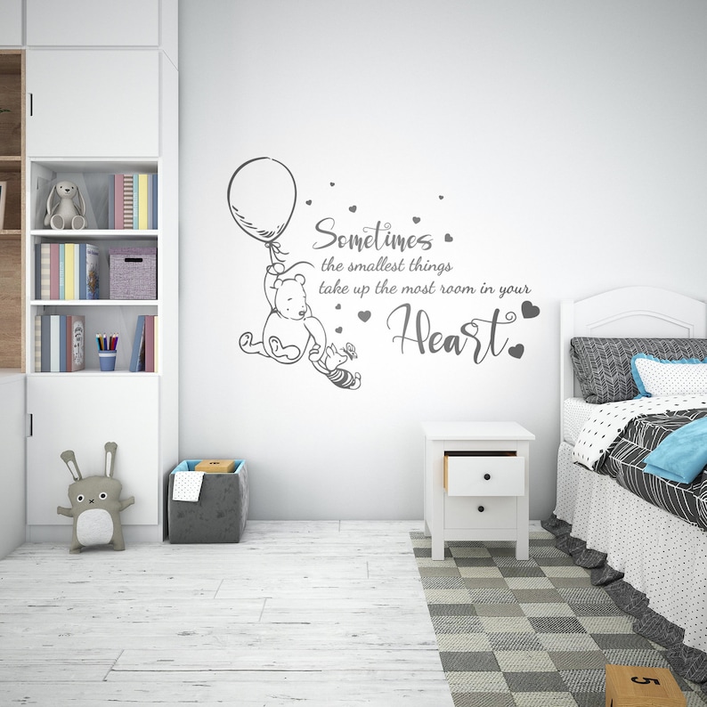 May include: A Winnie the Pooh wall decal with a quote that reads "Sometimes the smallest things take up the most room in your heart." The decal is in a grey colour and features Winnie the Pooh holding a balloon and Piglet flying behind him.